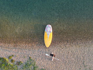Fototapeta premium Top-down drone view woman carrying paddle beside colorful board on pebbled lakeshore. Clear shallow water, warm sunlight, and graphic composition create calm sunny summer recreation scene
