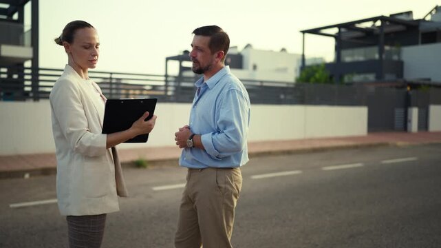 Professional female realtor with clipboard consulting male client about buying new home. Estate agent and  potential buyer stand on street in suburban area