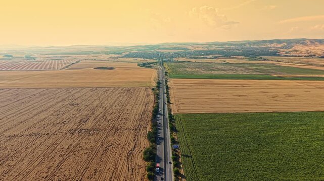 A long road runs through wide, open fields at sunset. The fields are separated by lines and have different colors. The sky is clear with some clouds.