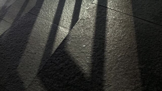 Parallel shadows stretch across stone pavement. Sunlight filters through vertical slats. Texture of stone contrasts with crisp, dark lines. No people, no animals, no plants present