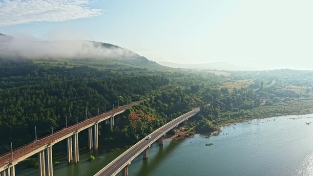 High-angle aerial view showing two parallel concrete highway bridges curving beside calm river shoreline with steady vehicle movement, soft morning fog, forested hills, and open scenic landscape