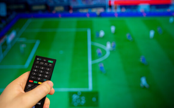 Hand holding TV remote control watching football match on screen.