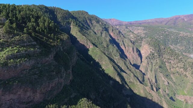 Aerial drone shot of lush green mountains and deep valleys on Tenerife. The drone pushes in, revealing rugged cliffs and a remote, forested landscape under a clear blue sky.
