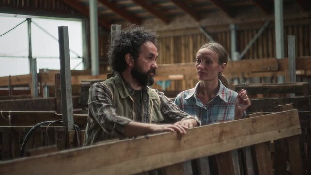 Male and female farmer in plaid shirts standing by wooden fence, discussing agricultural work tasks inside rustic livestock barn