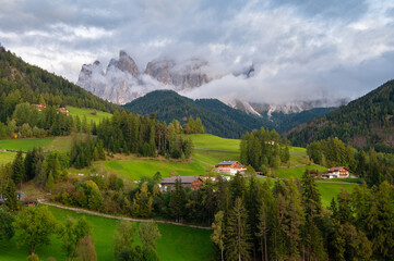 Fototapeta premium Traditional alpine chalets and misty Odle peaks in Val di Funes, Dolomites, Italy