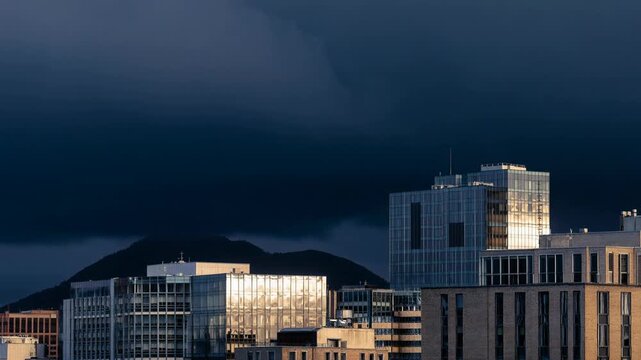 Modern cityscape with reflective glass buildings under dramatic stormy sky and mountain background