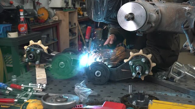 An engineer welds a frame, gear sprocket, and chassis for a homemade scooter/snowmobile with a small trailer.
