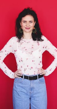Attractive playful Caucasian woman with hands on hips winking eye with flirty confident smile on bright red studio background, cheerful playful mood winking gesture stock promo concept video clip