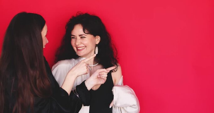 Two attractive Caucasian women pointing finger side showing direction with amazed surprised face and laughing on bright red studio background, excited friends promo discount concept stock video clip