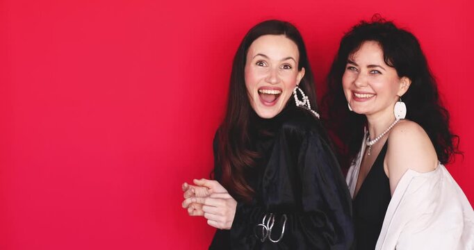 Two excited Caucasian women pointing fingers left side showing promo direction with shocked surprise face and laughing on bright red studio background, advertising deal concept stock video clip
