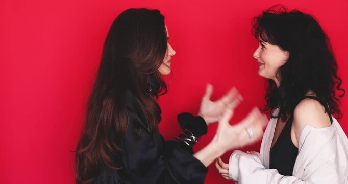 Two attractive Caucasian women standing side profile having emotional animated dialogue waving hand and pouting lips on bright red studio background, expressive discussion gesture stock video clip