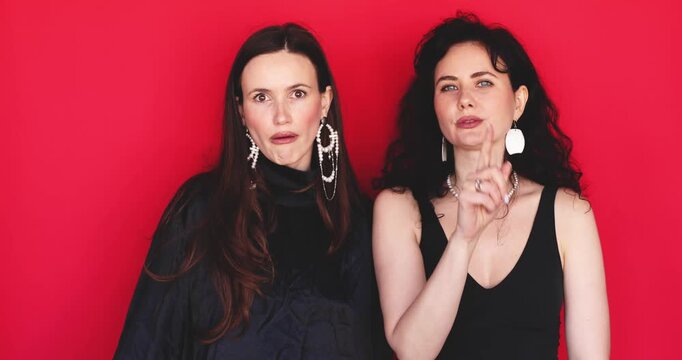 Two attractive Caucasian women putting index finger to lips showing shh gesture for silence and keeping secret looking at camera on bright red studio background, quiet moment stock video clip