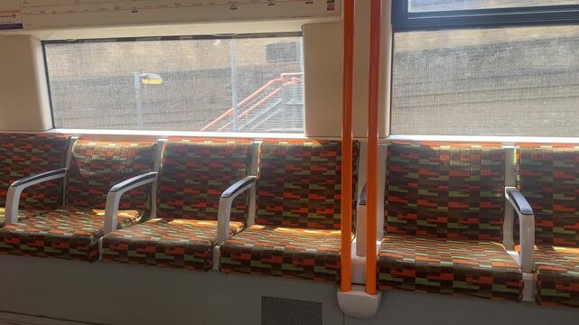 Interior view of a London Overground train featuring colorful moquette seats, longitudinal seating layout and orange handrails, captured during a passenger journey.