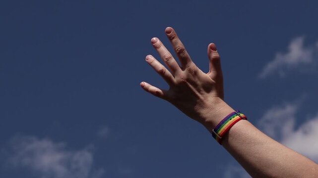 Close-up of a human hand with a bright rainbow textile bracelet (LGBTQ+ pride) on a sky background. Female hand raised to a clear blue sky. Freedom and equality concept