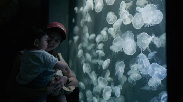 Happy Asian mother holding her cute baby boy watching illuminated jellyfish in aquarium.