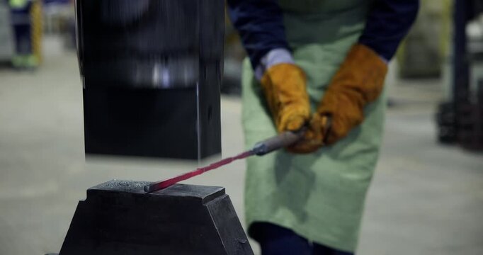Worker placing red hot metal rod on anvil in industrial workshop with protective gloves and apron.