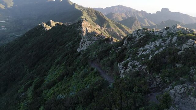 Aerial drone push in shot revealing the rugged mountain ridge of Cumbre del Carrizal on Tenerife, Canary Islands, at sunset. Lush vegetation, rocky peaks and a winding trail lead to a remote village.