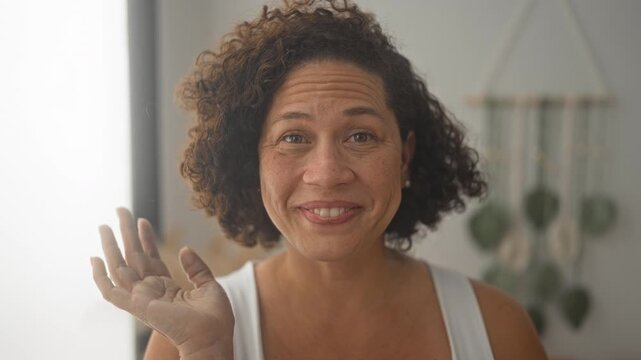 Woman middle aged hispanic smiles and waves hand at camera inside a building near hanging plants and window light; warmth connection.