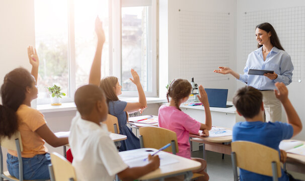 Diverse group of schoolchildren raising hands eagerly to answer teachers question during math class in sunlit classroom, encouraging participation and digital learning
