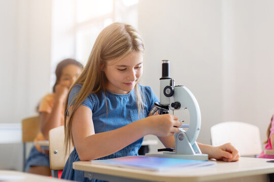 Focused schoolgirl adjusts a microscope during a science lesson, examining a specimen closely while classmates study in the background classroom