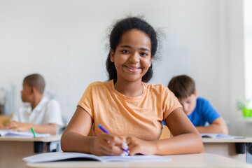Obraz premium Smiling black schoolgirl writing in open notebook at her desk and looking toward the camera, while diverse classmates study independently in a bright modern classroom behind her