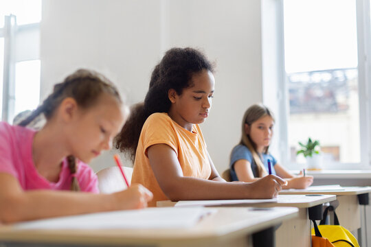 Diverse schoolgirls write assignments at individual desks during quiet classroom lesson, showing concentration, academic progress, inclusion, and independent learning
