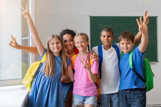 Five diverse schoolchildren posing together with colorful backpacks and cheerful hand gestures in bright classroom, expressing friendship, confidence, and school spirit
