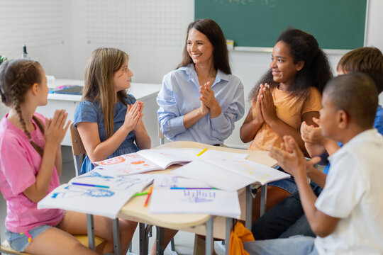 Smiling female teacher and diverse schoolchildren applaud after completing a creative classroom project, celebrating teamwork, achievement, participation, and shared academic success