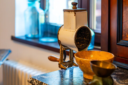 Vintage white enameled manual hand-crank grater for nuts and cheese mounted on a marble tabletop next to a ceramic bowl in a rustic kitchen near a window.