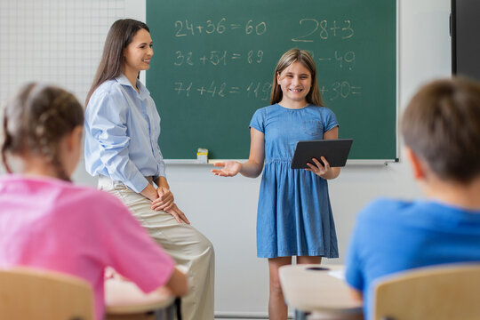 Confident schoolgirl using tablet to present mathematics work to classmates while her teacher offers support beside a chalkboard in a bright modern classroom