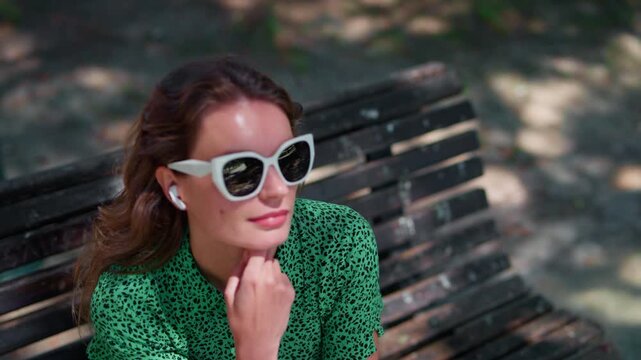 A woman sitting on a bench in summer, wearing stylish sunglasses and earbuds, pondering with a thoughtful expression against a blurred green background