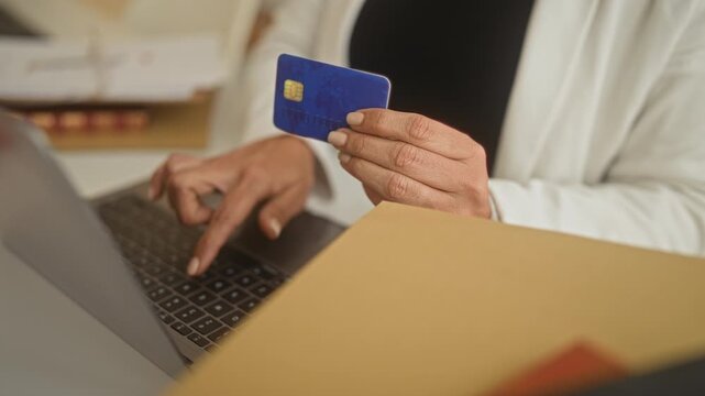 Woman holding blue creditcard with bare hand and typing on laptop inside building; online shopping focused.