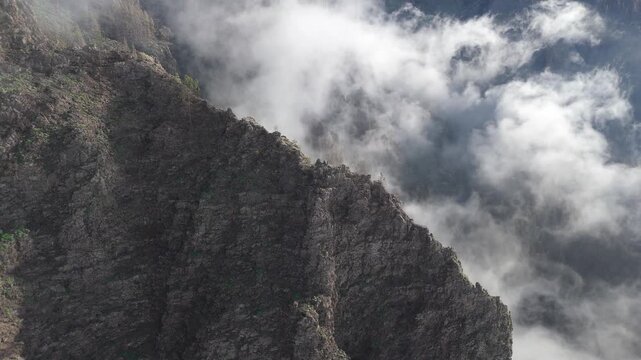 An aerial drone shot pushes in on the dramatic, fog-shrouded mountains near Morro Gallardo on the Tenerife, Canary Islands. Rugged cliffs and green vegetation emerge from thick clouds.