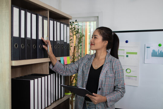 Young businesswoman searching through files in an archive, holding a clipboard while organizing documents and standing next to shelves filled with ring binders in a professional office.