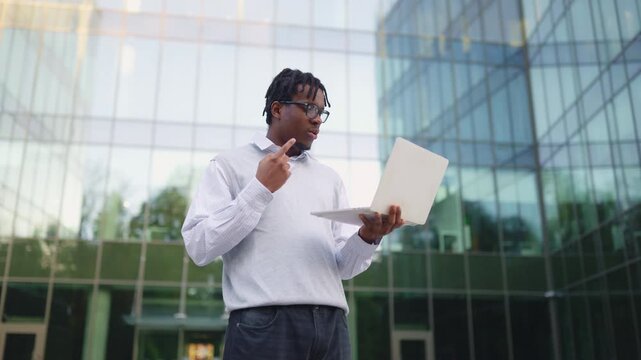 A young African American man with dreadlocks and glasses is holding a laptop and waving hello during a video call outdoors in front of a modern glass building