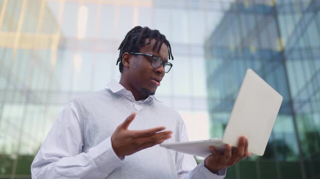 A smiling Black man with dreadlocks and glasses is holding a laptop and gesturing while on a video call outside a modern glass building