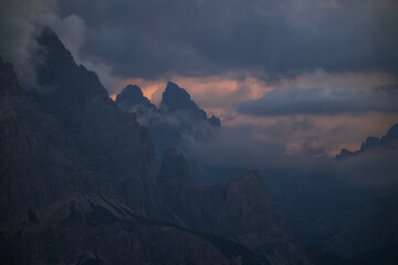 A dramatic landscape featuring jagged mountain summits partially covered by clouds and mist during a moody sunset or twilight. Ideal for themes of mystery, nature, and adventure. © marcodenaro