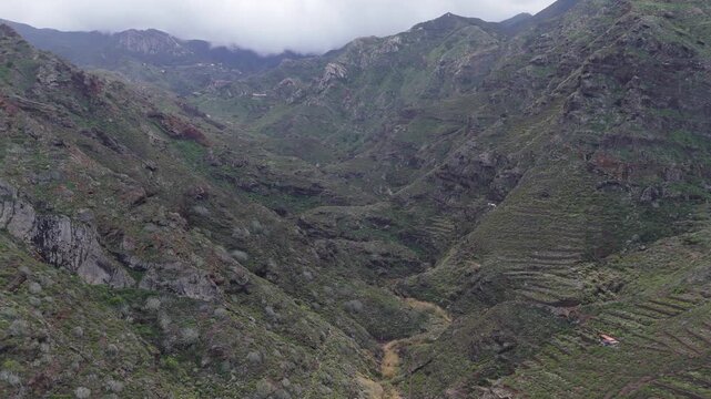 Push in and boom up aerial drone shot revealing a dramatic, green mountain valley with scattered houses on Tenerife. Cloudy skies highlight the rugged landscape of the Canary Islands.