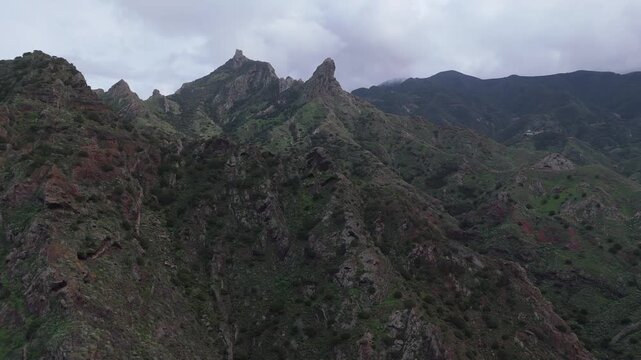 Aerial drone shot pushing in and booming up over the rugged, green mountain landscape of Roque de las &Aacute;nimas on Tenerife, Canary Islands. Capturing the remote, hilly terrain and cliffs.