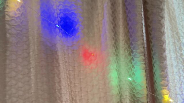 Colorful blinking christmas lights creating a festive bokeh effect behind a semi transparent textured curtain, casting red, blue, green, and yellow glows for a holiday mood