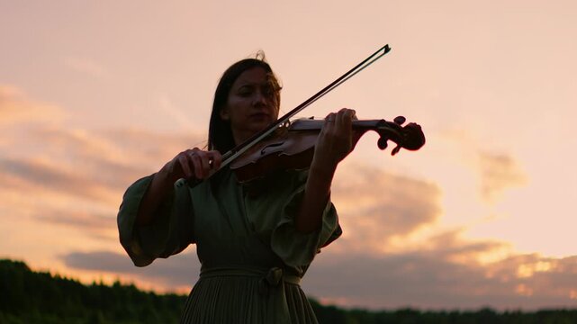 Woman playing violin outdoors at sunset, silhouetted against colorful sky with soft clouds, expressing emotion and serenity in a peaceful natural landscape scene