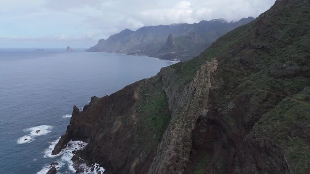 Aerial drone shot pushing in along the dramatic, rocky coast and lush green mountains on Tenerife, Canary Islands. Capturing the remote, untamed beauty of the Roque de las Animas mountain.