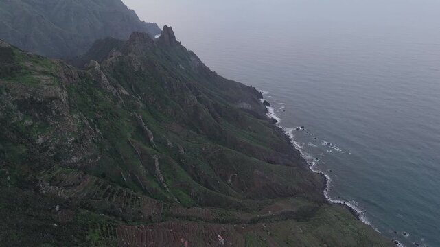 A dramatic aerial drone shot with a push-in movement revealing the rugged Anaga coastline and lush green mountains of Tenerife. Overcast sky above the expansive Atlantic Ocean.