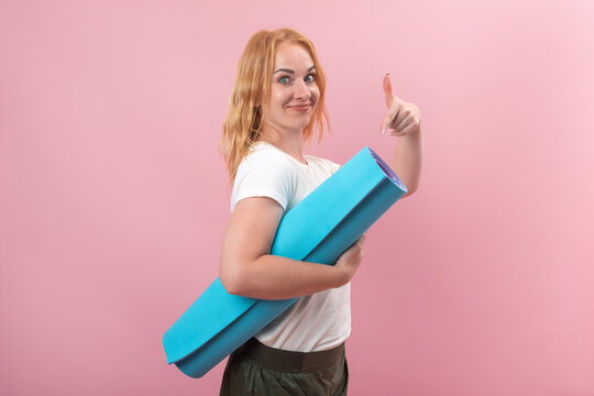 Young woman with red hair expressing approval and readiness for fitness activities, holding a blue yoga mat and showing a thumbs up gesture on a pink background