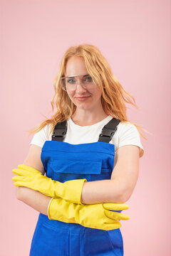 Young woman in protective workwear. Including blue overalls. Yellow gloves. And safety glasses. Standing confidently with crossed arms against a pink background