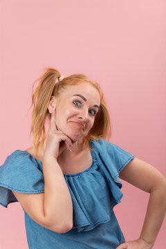 Young woman with red hair in pigtails is making a funny face while touching her chin, creating a playful and thoughtful expression on a pink background