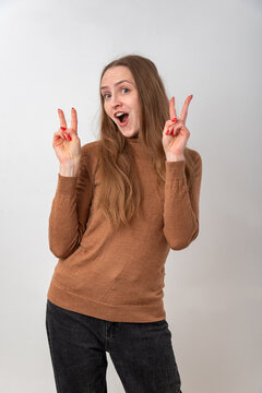 Young woman smiling with an excited expression, wearing a brown turtleneck and dark jeans, standing on a white background, making a V sign with both hands, representing success and happiness