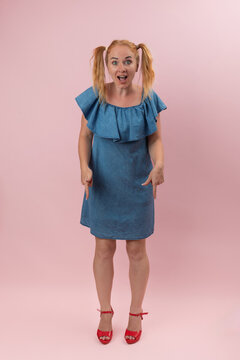 Young woman with blonde pigtails in a denim dress and red heels pointing down, expressing shock and amazement on a pink background