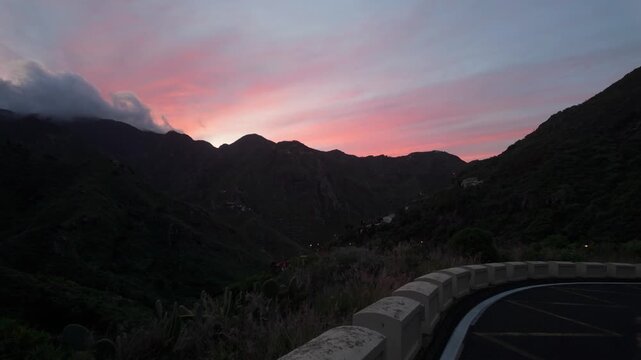 A breathtaking sunset paints the sky in vibrant pink and purple hues over the rugged, lush mountain valley of Anaga on Tenerife, Canary Islands. A winding road offers stunning views at dusk.