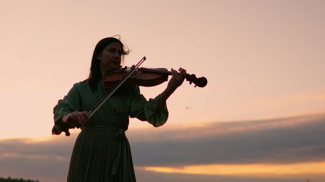 Woman playing violin outdoors at sunset with soft evening sky and gentle clouds in the background, creating a serene and peaceful musical silhouette scene